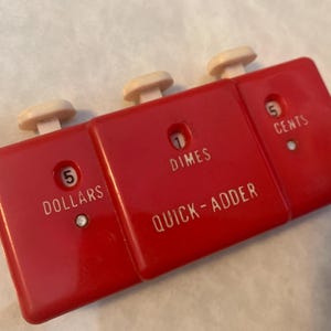 May include: A vintage red plastic Quick-Adder coin calculator with three sections. Each section has a white button and displays denominations: Dollars, Dimes, and Cents. The calculator is designed to quickly add up coins.