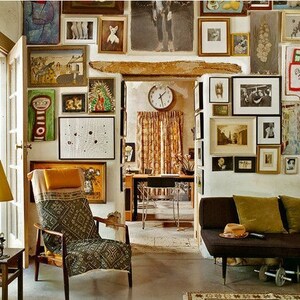 May include: A living room with a brown couch, a yellow lamp, and a chair with a patterned fabric. The walls are covered in framed artwork, including paintings, drawings, and photographs.