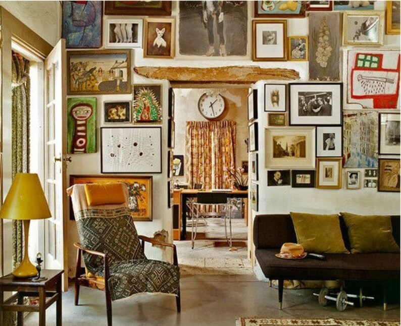May include: A living room with a brown couch, a yellow lamp, and a chair with a patterned fabric. The walls are covered in framed artwork, including paintings, drawings, and photographs.