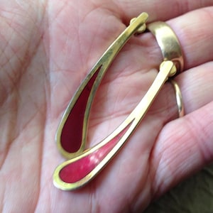 May include: Gold-toned earrings with a teardrop design. The earrings feature a red enamel inlay and a curved, elongated shape. The earrings are shown against a neutral background.