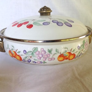 May include: White enamelware casserole dish with lid. The dish features a fruit design with red strawberries, purple plums, and orange peaches. It has gold-colored handles and a gold-colored rim.