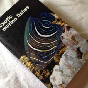 May include: Book cover featuring the title "exotic marine fishes" in white text on a black background. The cover image shows a colorful fish with blue and white stripes swimming in a coral reef.