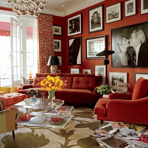 May include: A living room with red walls and a red velvet sofa, two red armchairs, a glass coffee table, and a yellow floral arrangement. The room is decorated with framed black and white photographs.