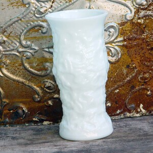 May include: A white milk glass vase with a textured, raised floral design. The vase is approximately 6 inches tall.