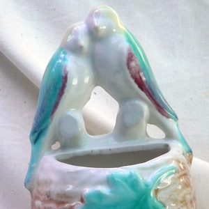 May include: A ceramic wall planter featuring two blue, green, and purple birds perched on a branch. The planter has a rectangular opening for planting.