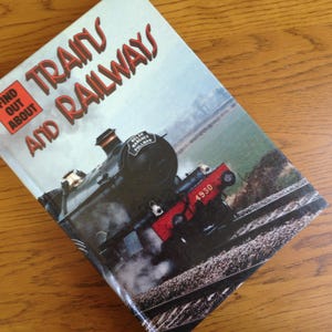 May include: A book titled "Trains and Railways" with a vintage train illustration on the cover. The cover features a black steam engine with red accents and the number 4930. The book is on a wooden surface.