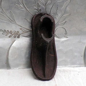 May include: A vintage, dark brown cast iron shoe last, a form used to shape shoes. The last is positioned vertically, showcasing its detailed form. The background features a silver metal surface with floral engravings.