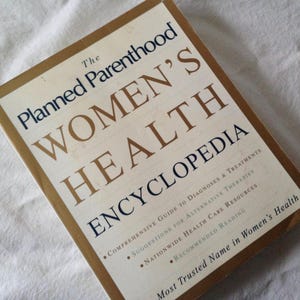 May include: A hardcover book titled "The Planned Parenthood Women's Health Encyclopedia." The cover is beige with gold lettering and a gold border. The book provides a comprehensive guide to diagnoses and treatments, suggestions for alternative therapies, and nationwide health care resources.