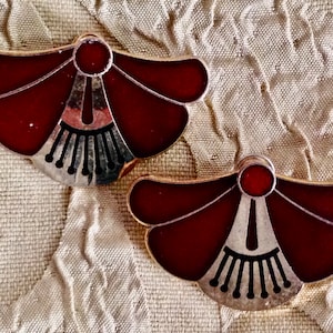 Pode incluir: Um par de brincos vintage com um design floral. Cada brinco apresenta uma forma de pétala de esmalte vermelho escuro com detalhes em metal prateado. Os brincos têm uma borda dourada e estão sobre um tecido bege texturizado.