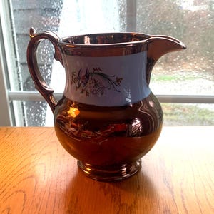May include: A copper-colored pitcher with a light blue band featuring floral designs. The pitcher has a rounded body, a handle, and a spout. It sits on a wooden surface near a window.
