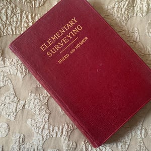 May include: A vintage, red hardcover book titled "Elementary Surveying" with gold lettering. The book's cover is slightly worn, and the title is above the author's names, Breed and Hosmer. The book is resting on a textured, off-white surface.
