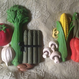 May include: A collection of colorful, sculpted vegetables including a red tomato, green celery, asparagus, corn, red bell pepper, and mushrooms. The vegetables are arranged on a textured, light-colored surface.