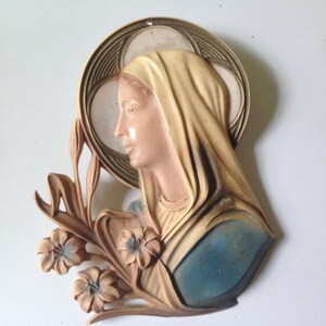 May include: A vintage wall hanging featuring a relief of a woman's profile, adorned with a halo and floral accents. The sculpture is painted in muted tones of beige, pink, and blue, creating a classic aesthetic. The piece is likely made of a composite material.