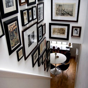 May include: A white wall with a gallery wall of black framed pictures and artwork. The pictures are of various sizes and shapes and include photos, drawings, and prints. The wall is in a home with a wooden staircase and a white table with black chairs.