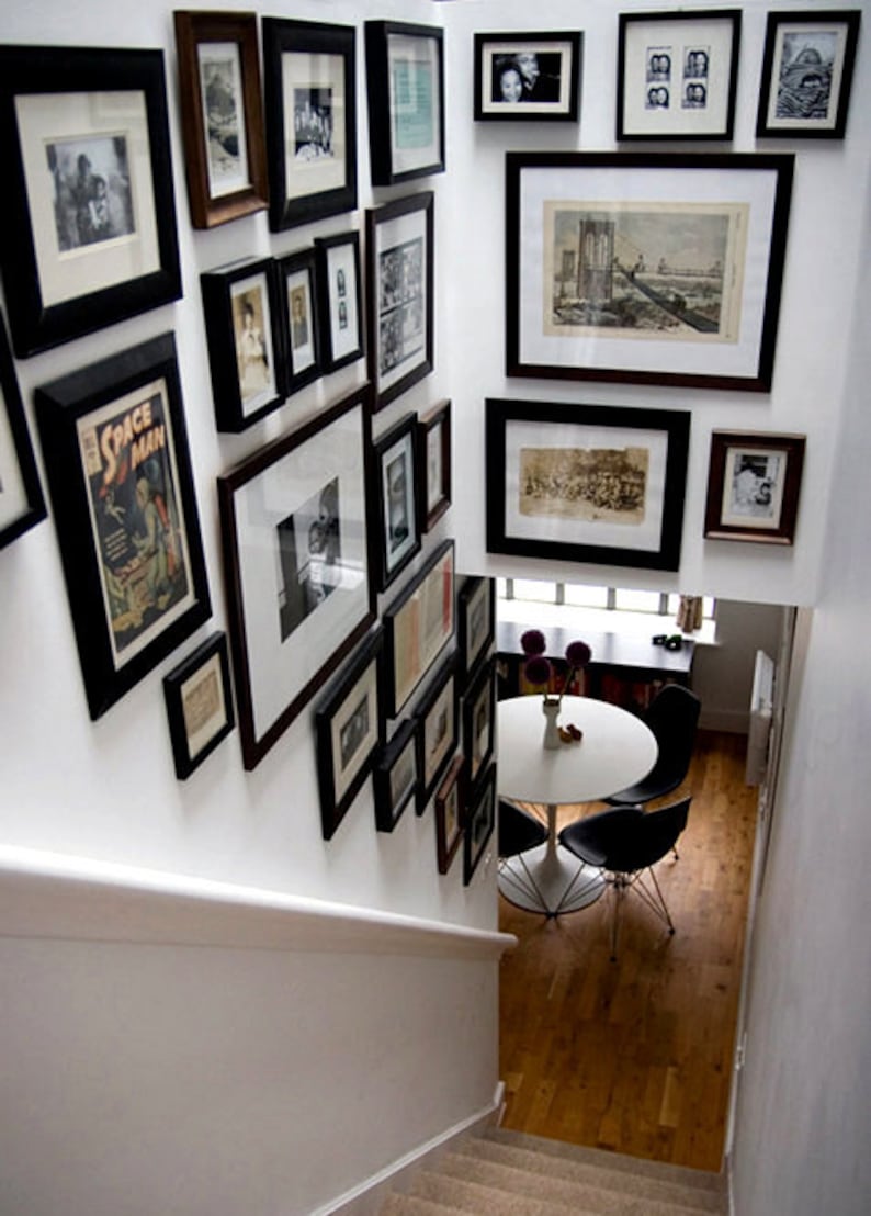 May include: A white wall with a gallery wall of black framed pictures and artwork. The pictures are of various sizes and shapes and include photos, drawings, and prints. The wall is in a home with a wooden staircase and a white table with black chairs.
