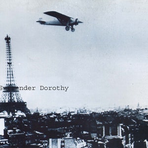 May include: A black and white photo of the Eiffel Tower in Paris, France. A small airplane flies over the tower. The text "Surrender Dorothy" is printed on the image.