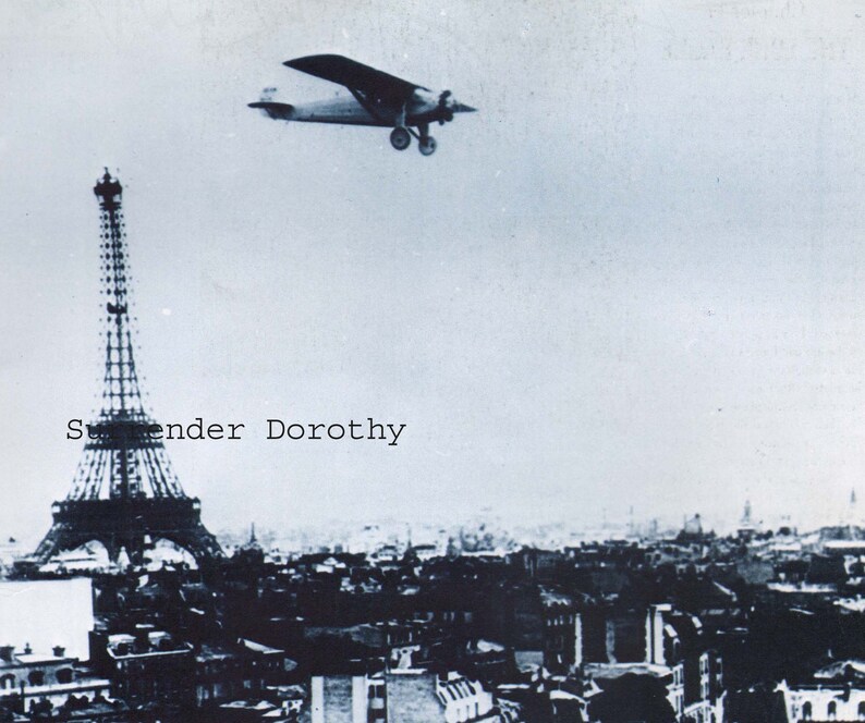 May include: A black and white photo of the Eiffel Tower in Paris, France. A small airplane flies over the tower. The text "Surrender Dorothy" is printed on the image.