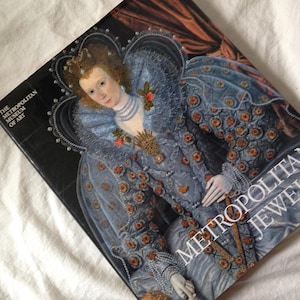 May include: A book titled "Metropolitan Jewelry" from The Metropolitan Museum of Art. The cover features a portrait of a woman in a blue gown with intricate details and jewelry. The background is a soft, neutral color.