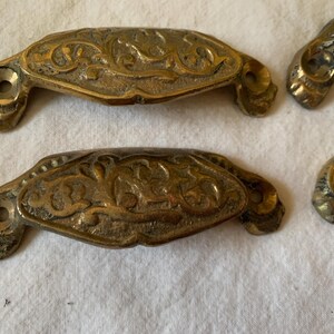 May include: Close-up of several antique brass drawer pulls. The pulls have an ornate, floral design and are a warm, golden color. Each pull has two screw holes for mounting. The pulls are likely vintage and add a touch of elegance to furniture.