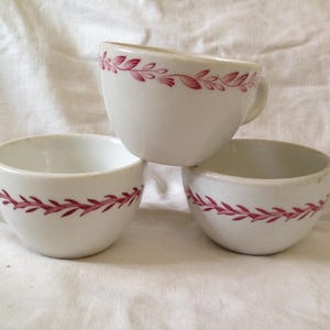 May include: Three white ceramic teacups with a pink floral design around the rim.