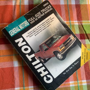 May include: A Chilton repair manual for General Motors full-size trucks, covering 1988-98 models. The cover features a red pickup truck and includes wiring and vacuum diagrams. The manual is on a plaid orange, red, and yellow surface.