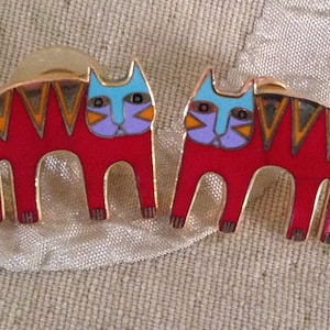 May include: A pair of red cat-shaped earrings with blue and purple details. The cats have a geometric pattern on their backs and gold accents.