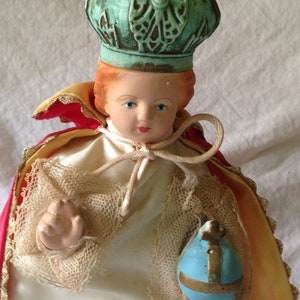 May include: A small porcelain doll dressed in a white gown with lace trim, a red and gold cape, and a blue and gold crown. The doll is holding a blue and gold orb.