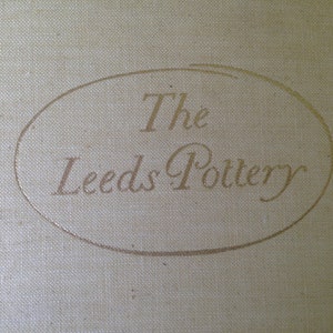 May include: A close-up of a white fabric surface with a gold oval logo that reads "The Leeds Pottery".