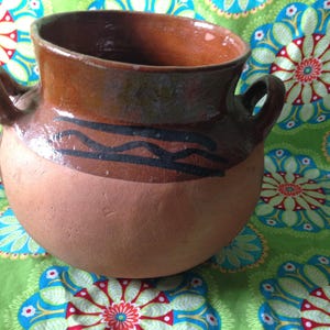 May include: A brown clay pot with two handles and a black and brown wavy design around the middle. The pot is sitting on a green and blue floral fabric.