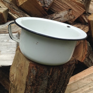 May include: White enamel chamber pot with a black rim and a handle. The pot is sitting on a wooden stump.
