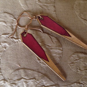 Puede incluir: Un par de pendientes de oro con un diseño geométrico rojo y dorado. Los pendientes son largos y estrechos, con una punta puntiaguda.