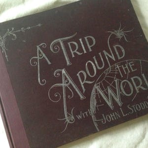 May include: A burgundy book with silver lettering that reads "A Trip Around the World with John L. Stoddard".