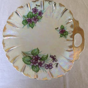 May include: A vintage, scalloped-edge dish with a pearlescent finish and gold trim. The dish is decorated with hand-painted purple violets and green leaves. It has a handle on one side, perfect for serving small treats.