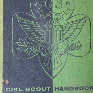 May include: A vintage Girl Scout Handbook with a green and blue cover featuring the Girl Scout emblem, a stylized eagle with wings spread, holding a branch in one claw and arrows in the other. The text "GIRL SCOUT HANDBOOK" is printed in black at the bottom of the cover.