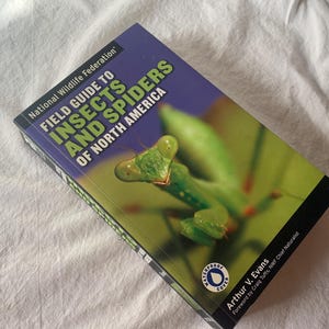 May include: A field guide titled "Field Guide to Insects and Spiders of North America" by Arthur V. Evans. The cover features a close-up of a green praying mantis against a blurred green background. The book has a blue and black spine.