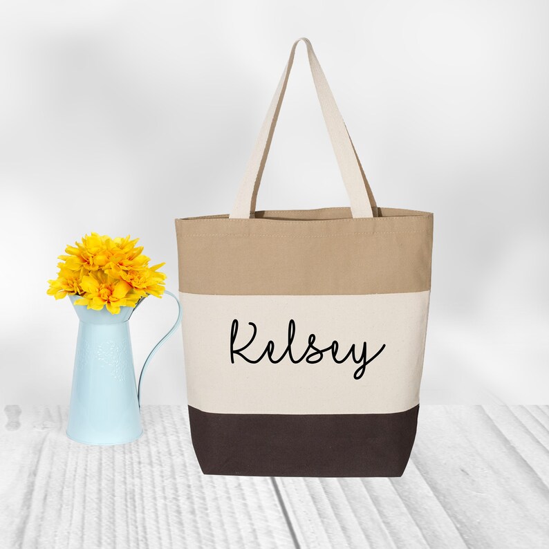 May include: A beige, white, and brown canvas tote bag with the name "Kelsey" printed in black on the front.