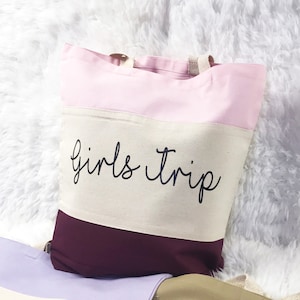 May include: A pink, beige, and burgundy striped tote bag with the text "girls trip" in black cursive lettering.