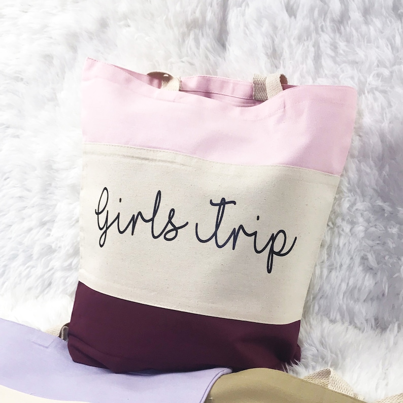 May include: A pink, beige, and burgundy striped tote bag with the text "girls trip" in black cursive lettering.