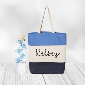 May include: A blue, white, and navy blue striped canvas tote bag with the name "Kelsey" embroidered in black on the front.