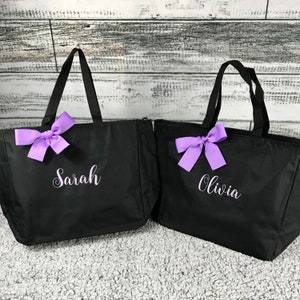 K&ouml;nnte beinhalten: Zwei schwarze Tragetaschen mit schwarzen Griffen und lila Schleifen. Eine Tasche hat den Namen "Sarah" in wei&szlig;er Schrift, die andere "Olivia". Die Taschen stehen auf einer hellgrauen Oberfl&auml;che mit wei&szlig;em Holzhintergrund.