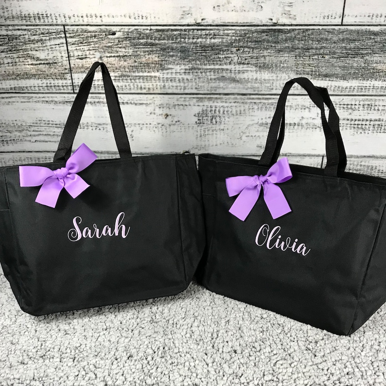 K&ouml;nnte beinhalten: Zwei schwarze Tragetaschen mit schwarzen Griffen und lila Schleifen. Eine Tasche hat den Namen "Sarah" in wei&szlig;er Schrift, die andere "Olivia". Die Taschen stehen auf einer hellgrauen Oberfl&auml;che mit wei&szlig;em Holzhintergrund.