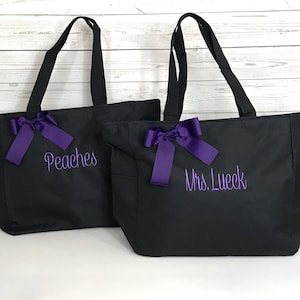 May include: Two black tote bags with purple embroidered text and purple bows. One bag says "Peaches" and the other says "Mrs. Lueck". The bags have black handles and are likely made of a durable fabric.