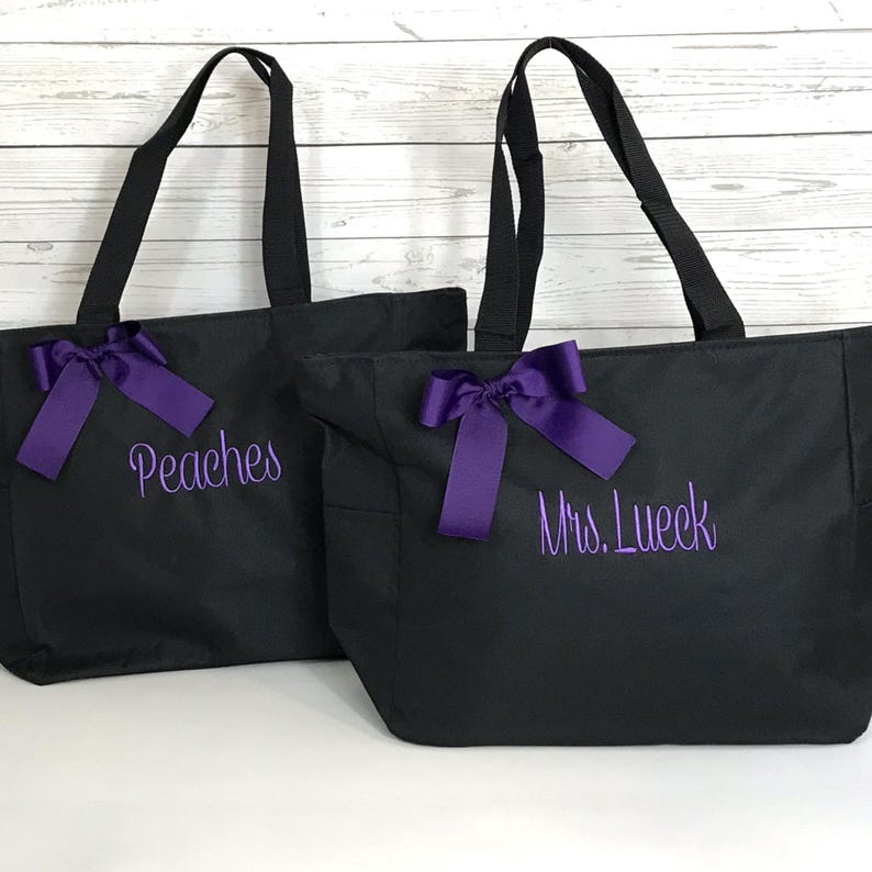 May include: Two black tote bags with purple embroidered text and purple bows. One bag says "Peaches" and the other says "Mrs. Lueck". The bags have black handles and are likely made of a durable fabric.