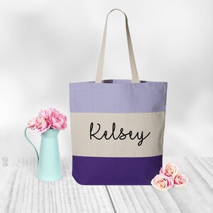 May include: A purple, beige, and white striped canvas tote bag with the name "Kelsey" printed in black on the beige stripe.