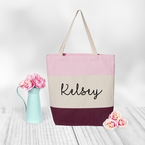 May include: A pink, beige, and burgundy canvas tote bag with the name "Kelsey" printed in black on the beige section.