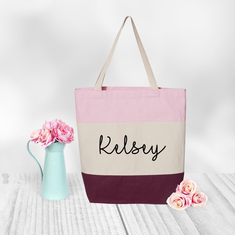 May include: A pink, beige, and burgundy canvas tote bag with the name "Kelsey" printed in black on the beige section.