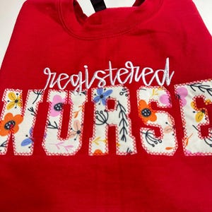 May include: A red sweatshirt with the words "registered NURSE" embroidered in white script and floral fabric letters. The floral fabric features a variety of colourful flowers and leaves. The sweatshirt has a crew neck and a black tag.