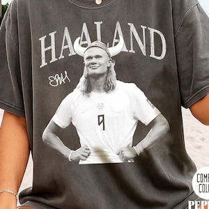 May include: A charcoal gray short-sleeve t-shirt featuring a graphic of a soccer player wearing a Viking helmet. The text HAALAND is printed above the image in a distressed white font, accompanied by a signature and the number 9 on the jersey.
