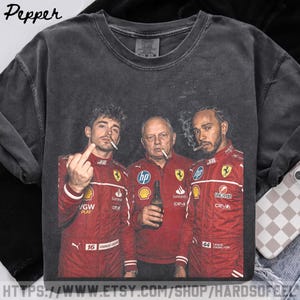 Pode incluir: Uma t-shirt de gola redonda cinzento-carvão com uma impressão gráfica de três pessoas com casacos de corrida vermelhos. A figura central segura uma garrafa, enquanto as figuras dos lados seguram cigarros. A figura da esquerda faz um gesto com o dedo médio.