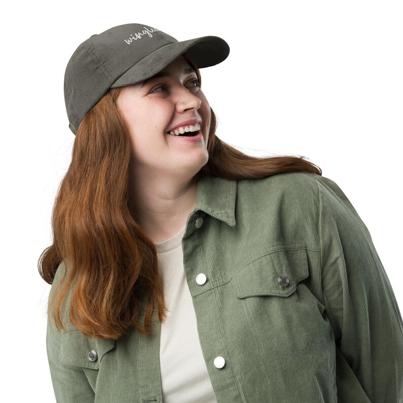 Pu&ograve; includere: Una donna indossa una giacca di velluto a coste verde e un berretto da baseball grigio scuro con la parola "wingless" ricamata. Sta sorridendo e guardando a destra.